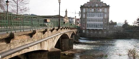 A stone bridge with a metal railing over a river, leading to a historic building with multiple windows and a clock tower.