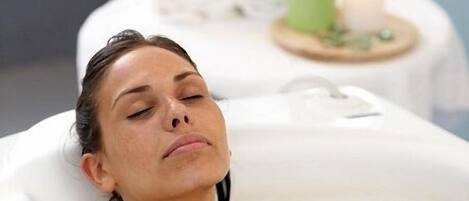 A person in a bathtub with rose petals floating on the water, a towel, and a candle on a side table in the background.
