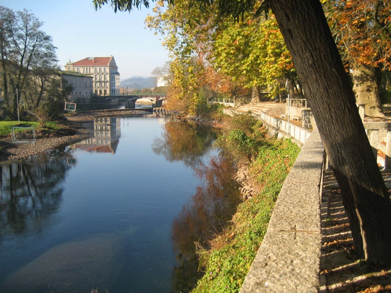 A calm river with a stone bridge, a building with a red roof, and a tree-lined path.