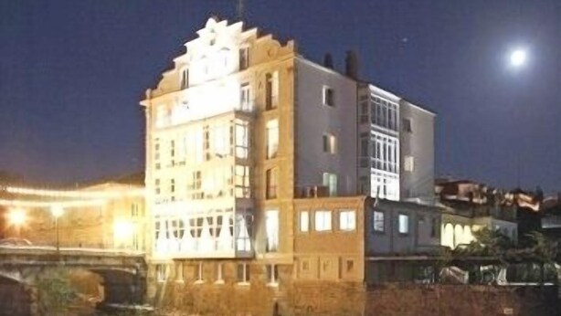 A multi-story building with a flat roof and a rectangular shape, situated next to a river with a bridge in the background. - Balneario Acuña (Caldas de Reis)