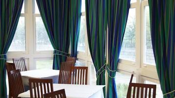 Un comedor con mesas y sillas, ventanas amplias con cortinas y vistas al exterior.