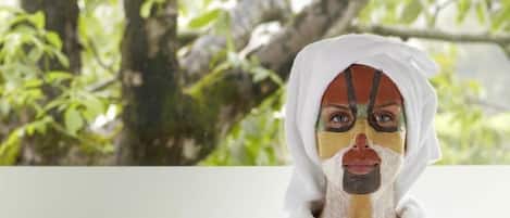 Una persona con bata blanca sentada junto a una ventana con vistas a árboles, rodeada de productos de spa y velas.