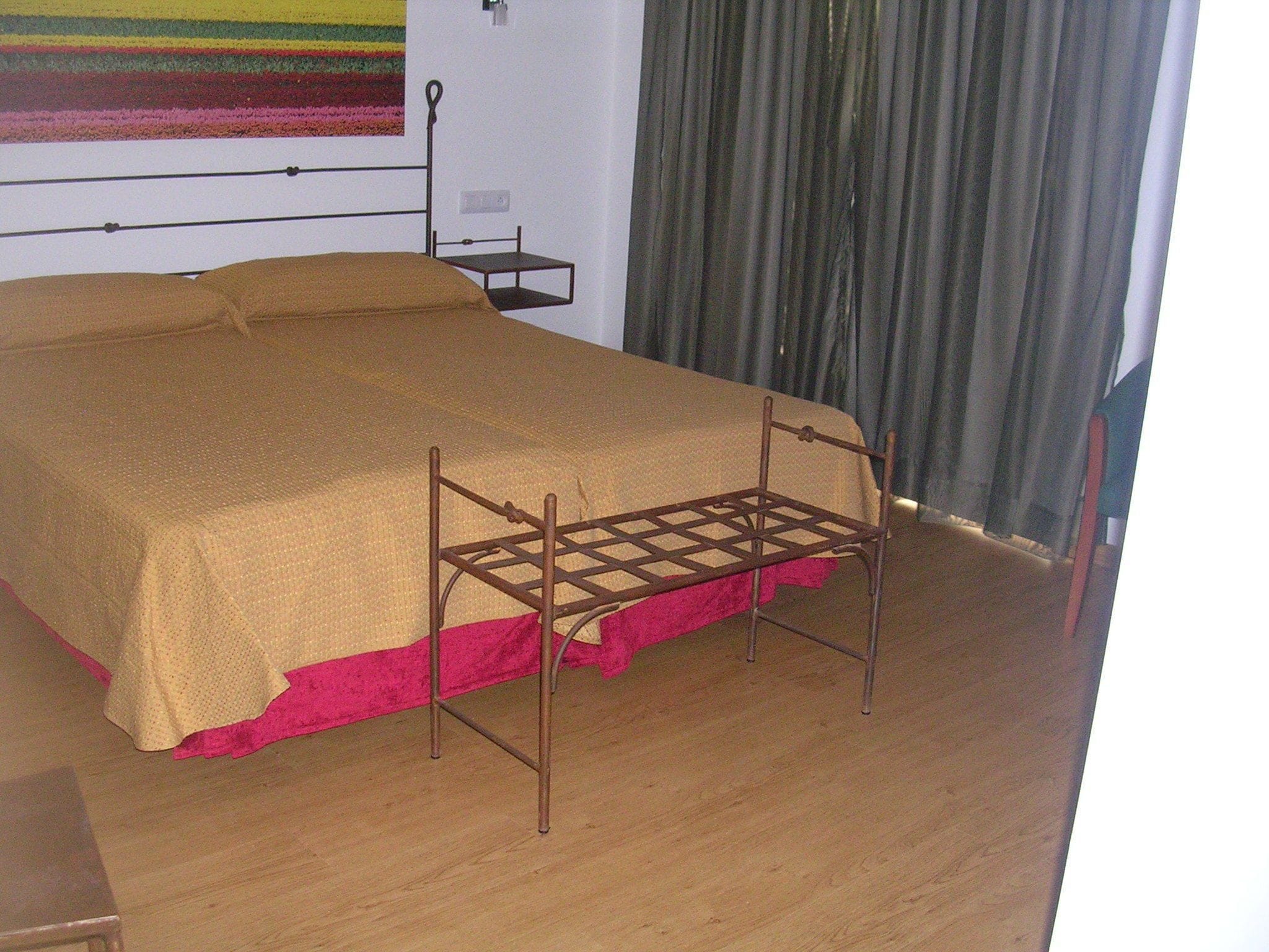 Una cama con estructura metálica y falda rosa, suelo de madera y pared a rayas.