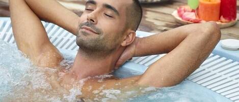 A person relaxing in a hot tub with candles and fruits on the side.