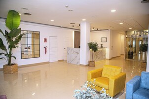 Un moderno vestíbulo de hotel con un mostrador de recepción, un sillón amarillo, un sillón azul y una planta grande en maceta.