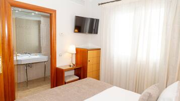 A hotel room with a bed, a TV mounted on the wall, a bathroom with a sink and mirror, and a wooden cabinet.