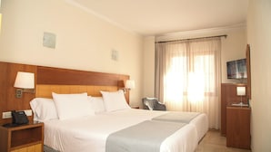 Habitación de hotel con una cama grande, una mesita de noche con teléfono, un escritorio con televisor y una ventana con cortinas.