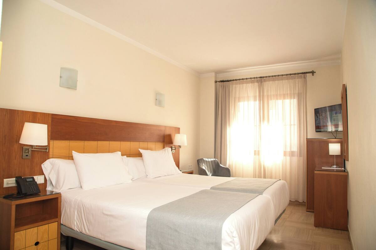 Une chambre d’hôtel avec un grand lit, une table de chevet avec un téléphone, un bureau avec une télévision et une fenêtre avec des rideaux.