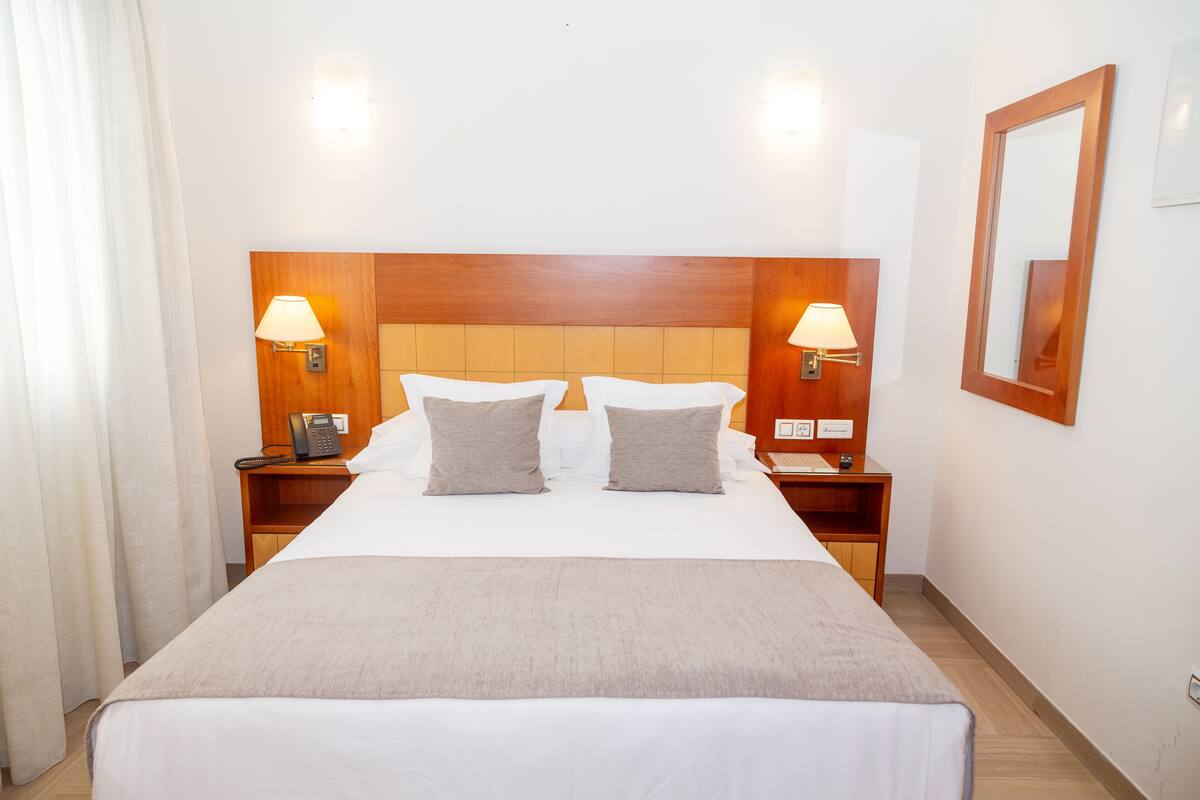 Une chambre d’hôtel avec un grand lit, deux tables de chevet avec des lampes, une tête de lit en bois et un miroir au mur.