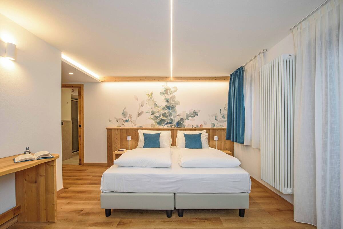A modern bedroom with a double bed, bedside tables, a nightstand, a wooden desk, and a wall-mounted radiator.