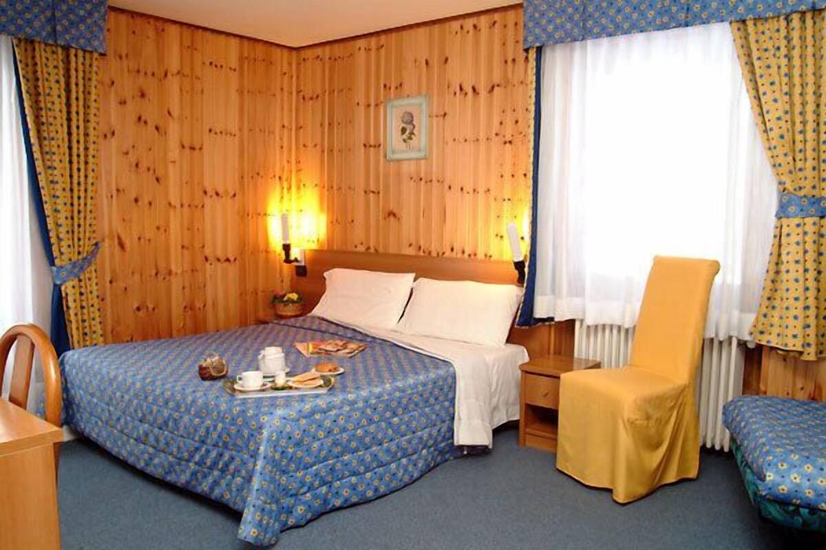 A hotel room with a wooden paneled wall, a bed with a blue floral bedspread, a yellow armchair, and a small table with a teapot and cups.