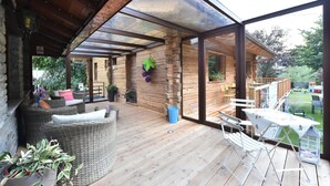 Un patio techado con mobiliario de mimbre, una mesa y sillas, con vistas a un jardín que incluye una terraza de madera y una zona de juegos.