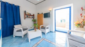 A room with a blue curtain, white wicker furniture, a television, a bookshelf, and a potted plant. - Hotel Celeste (Procida)