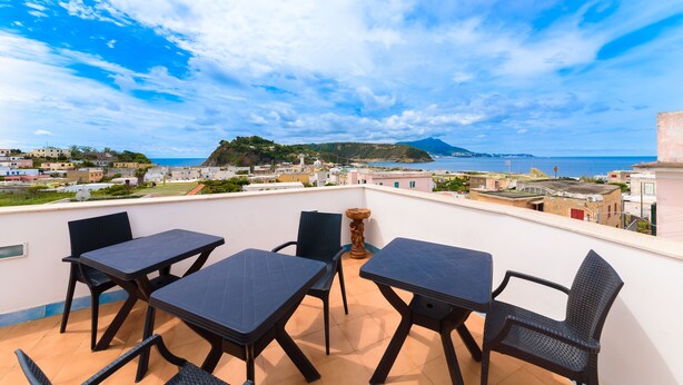 A terrace with four dark tables and chairs, overlooking a coastal town and the sea. - Hotel Celeste (Procida)