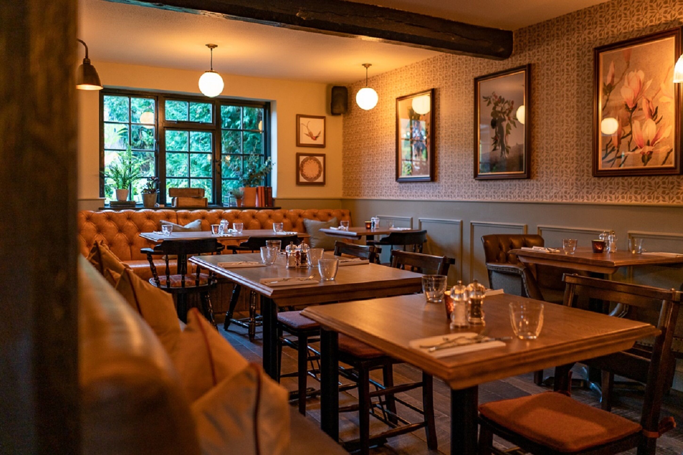 A restaurant with wooden tables and benches, framed artwork on the walls, and large windows with a view of greenery.