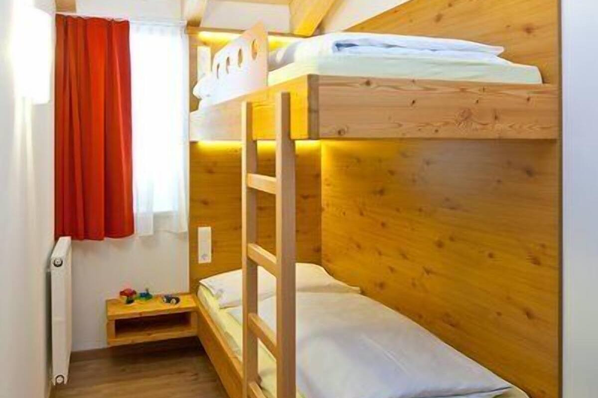 A bunk bed room with wooden walls and floors, a small table with toys, and a window with red curtains.