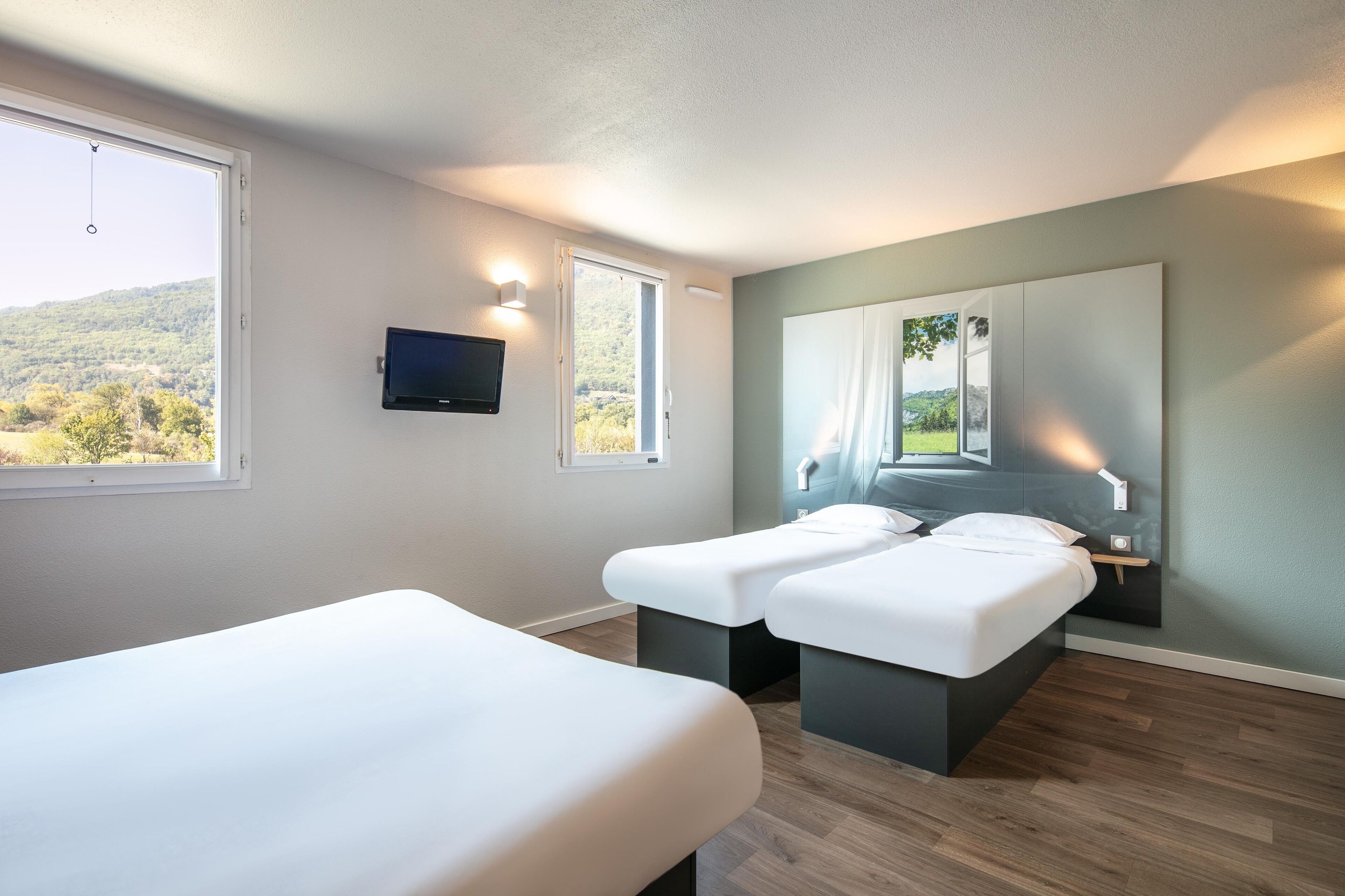 A modern hotel room with two beds, a large window with a view of a forest, a mounted TV, and a mirror reflecting the window view.