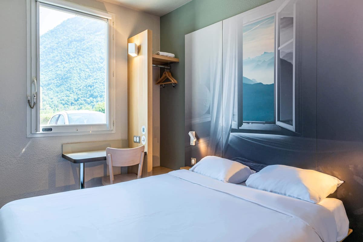 Ein Hotelzimmer mit Bett, Schreibtisch, Stuhl, Fenster mit Bergblick und einem an der Wand befestigten Ablagefach mit einem Handtuch.