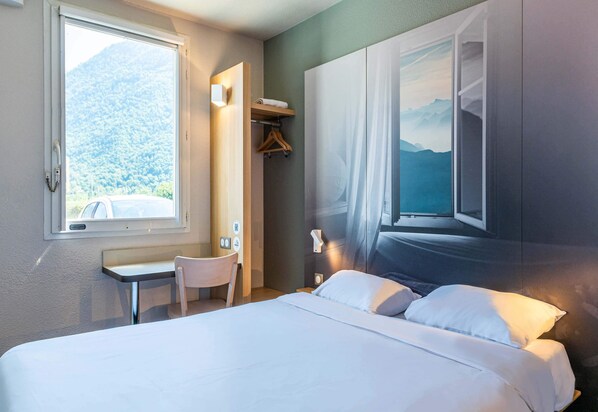 A hotel room with a bed, a desk, a chair, a window with a mountain view, and a wall-mounted shelf with a towel.
