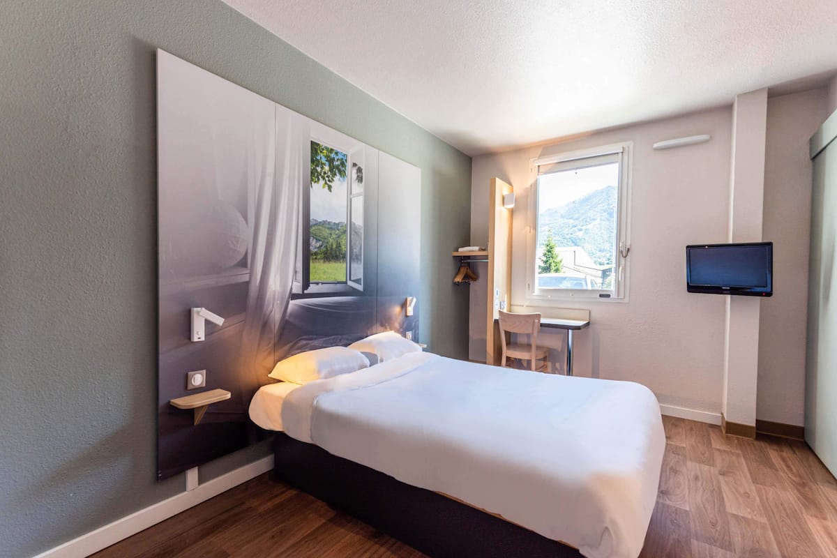 Ein modernes Hotelzimmer mit einem großen Bett, einem Schreibtisch, einem Sessel, einem Fernseher und einem Fenster mit Bergblick.