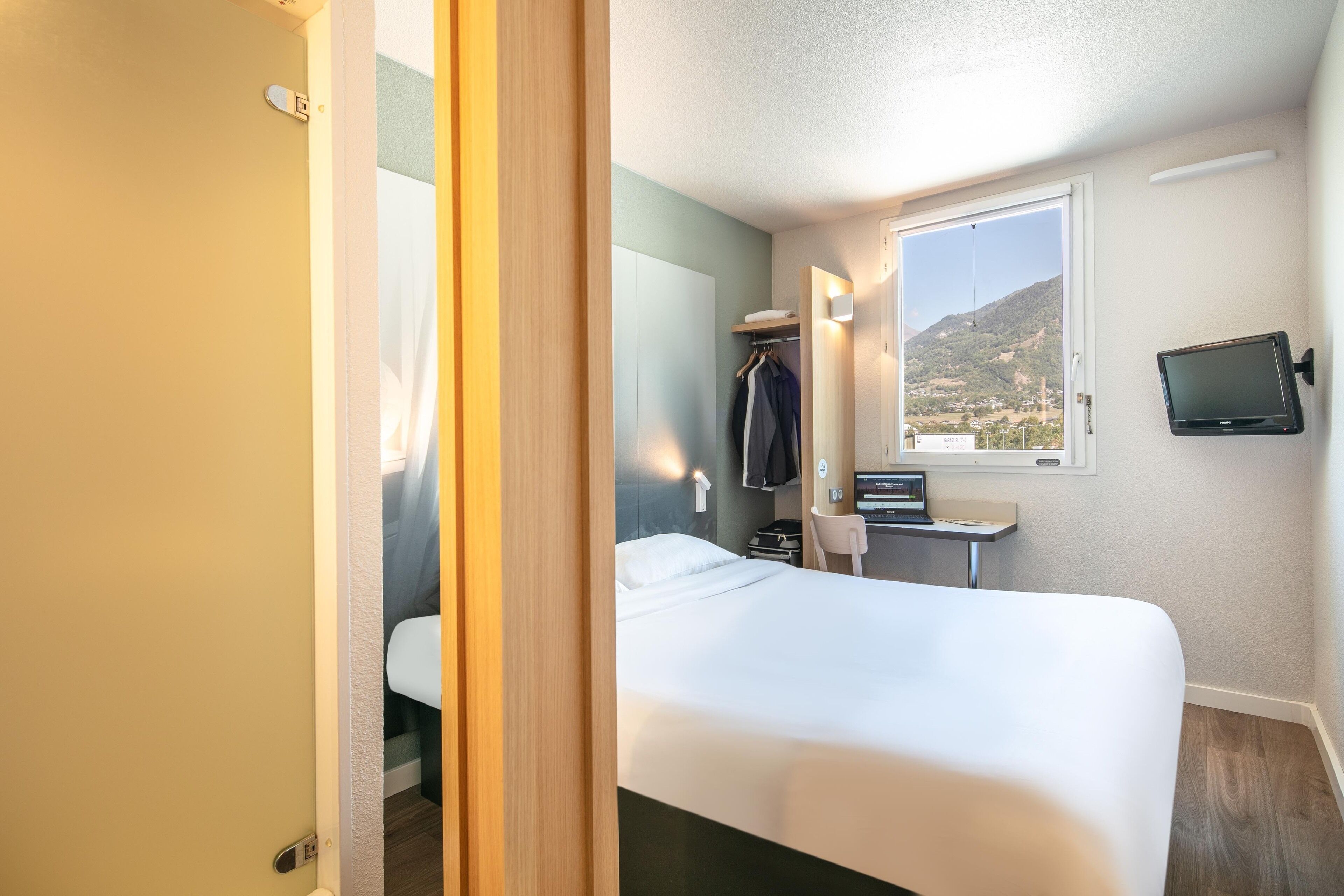 A hotel room with a bed, a desk with a computer, a mirror, and a window with a view of a mountain.