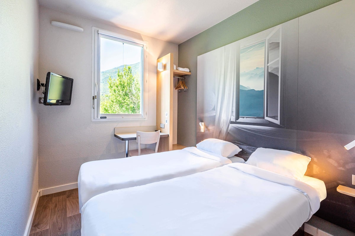 Ein Hotelzimmer mit zwei Betten, einem Fernseher, einem Fenster mit Blick auf Grünflächen und einem Bild an der Wand.