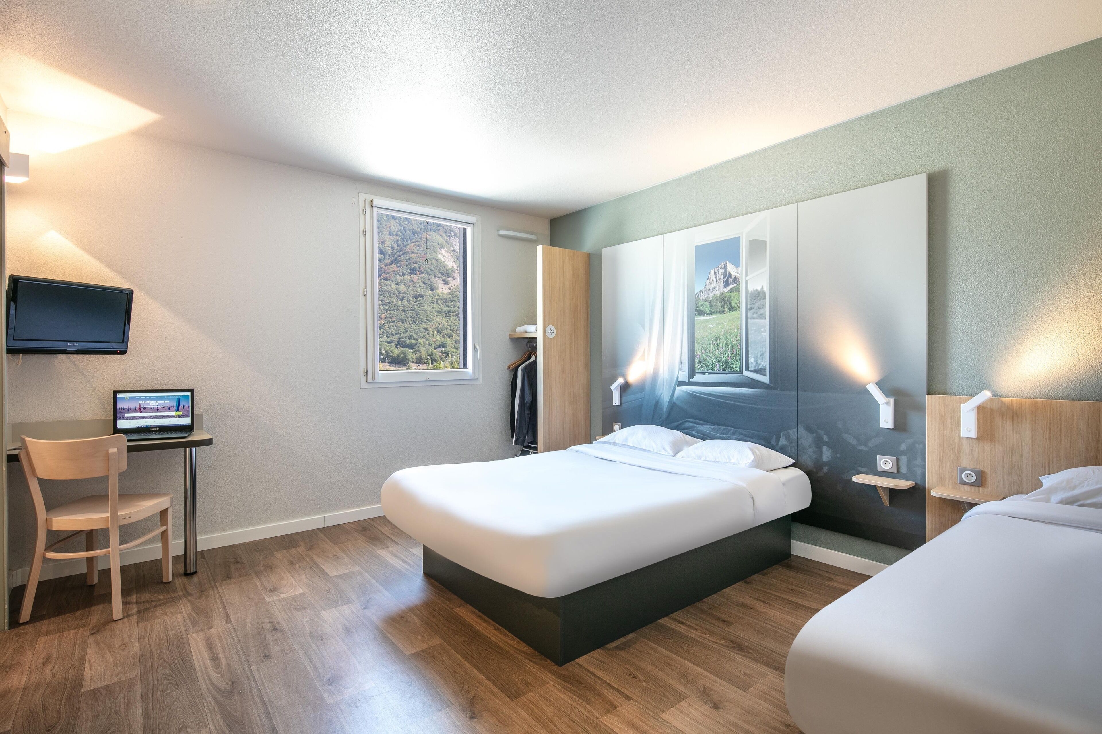 A modern hotel room with a large bed, a desk with a laptop, and a TV mounted on the wall.