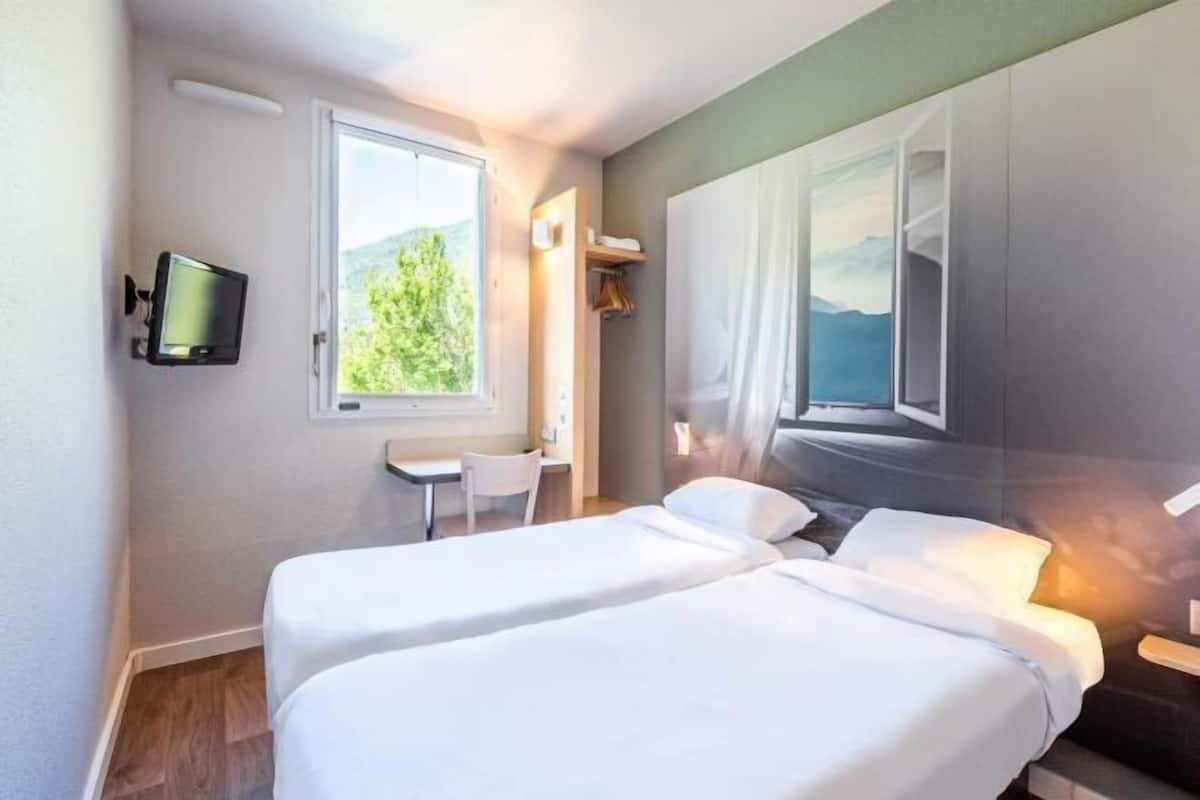 Ein Hotelzimmer mit zwei Betten, einem Fernseher, einem Fenster mit Ausblick und einem kleinen Tisch.