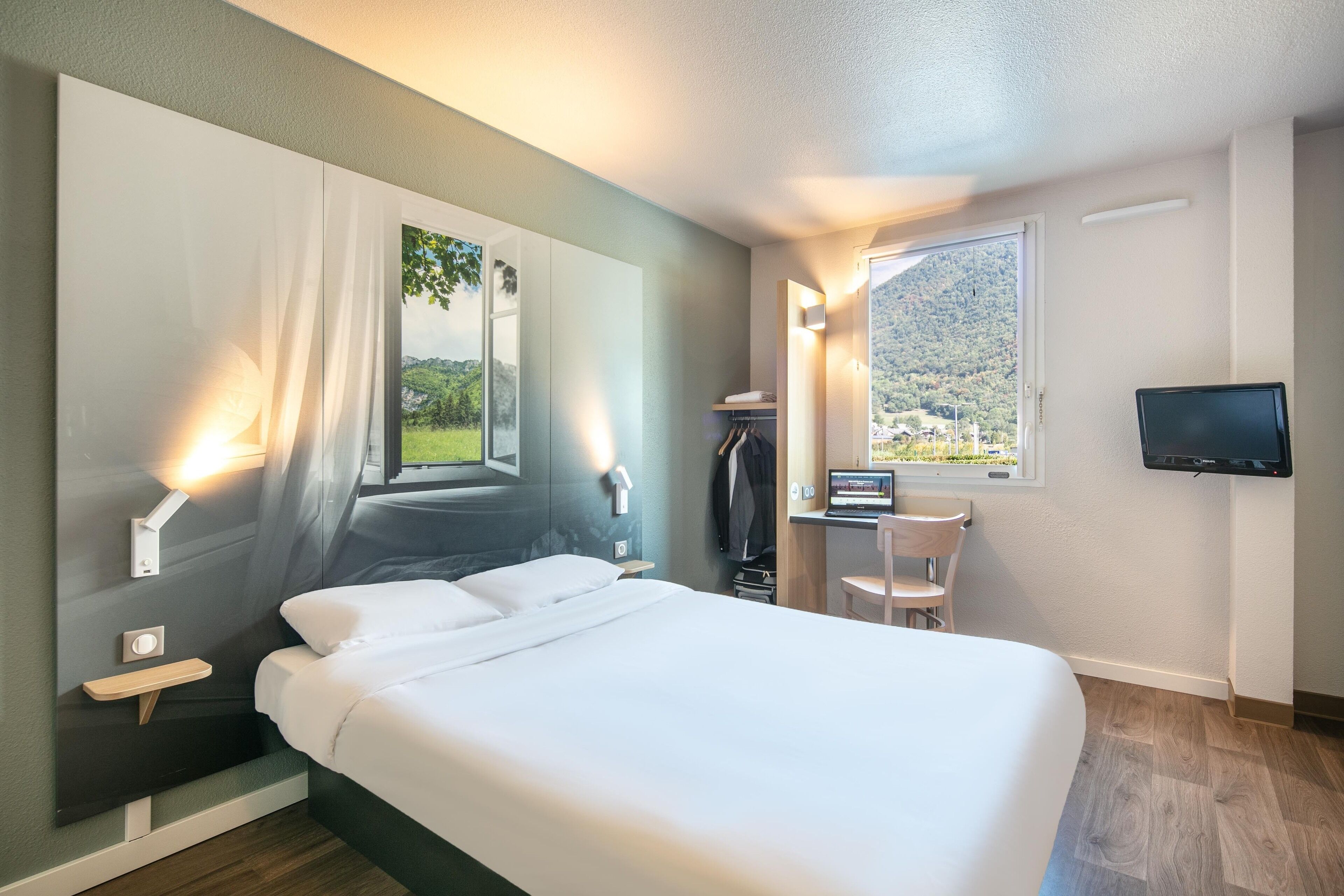 Une chambre d’hôtel moderne dotée d’un grand lit, d’un bureau avec une chaise, d’une télévision et offrant une vue sur un paysage montagneux.