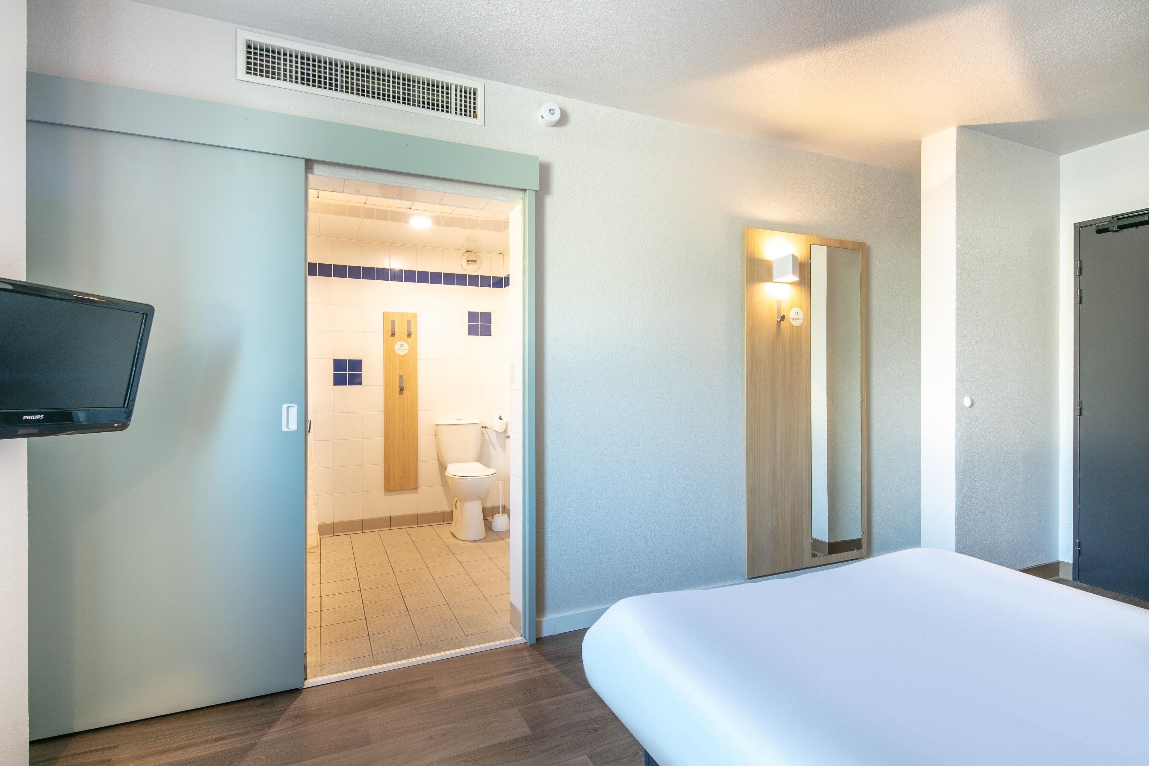 Quarto de hotel com cama, televisão, banheiro com vaso sanitário e pia, e espelho.