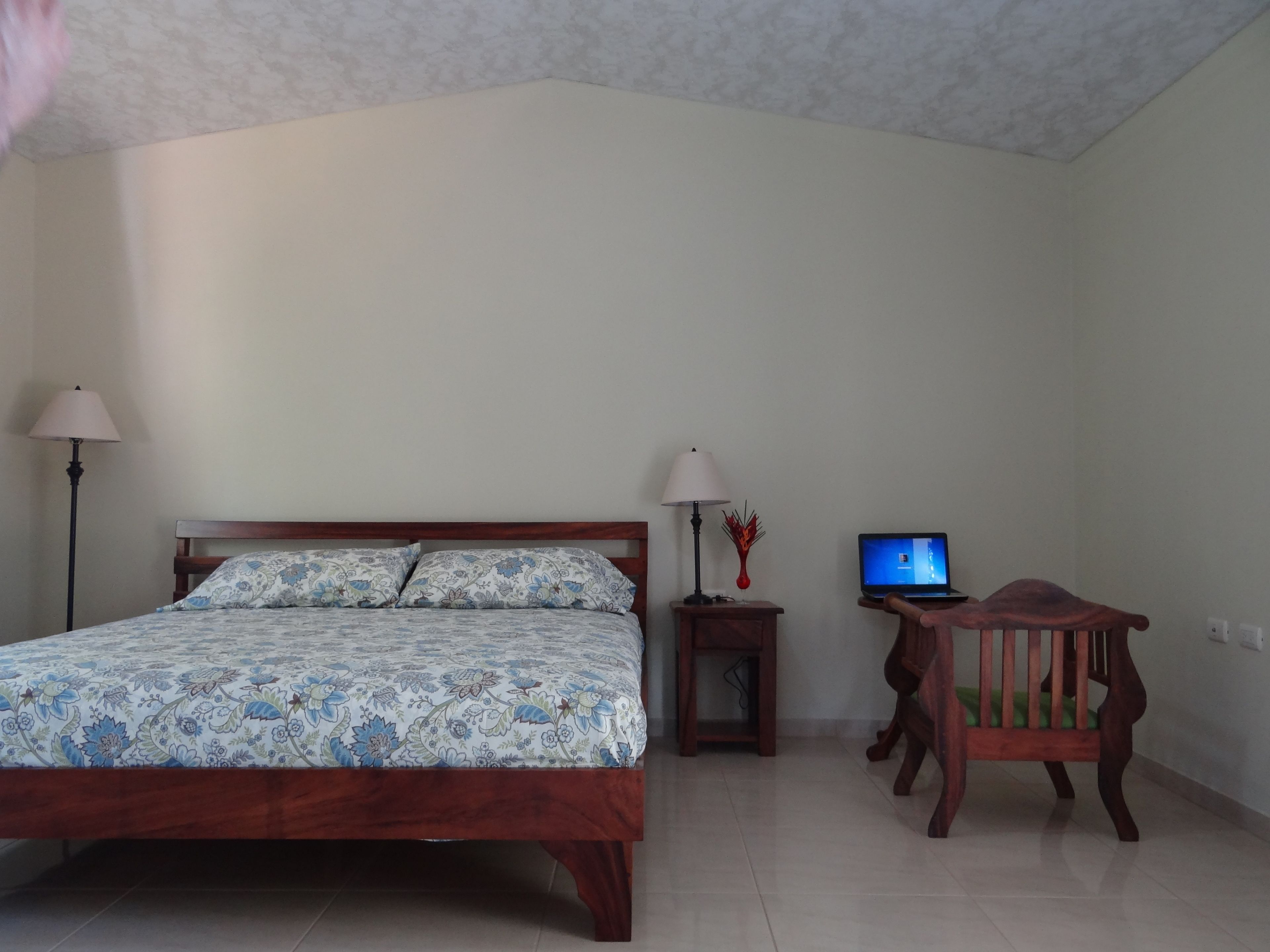 A bedroom with a bed, two bedside lamps, a small wooden nightstand with a laptop, and a wooden chair.