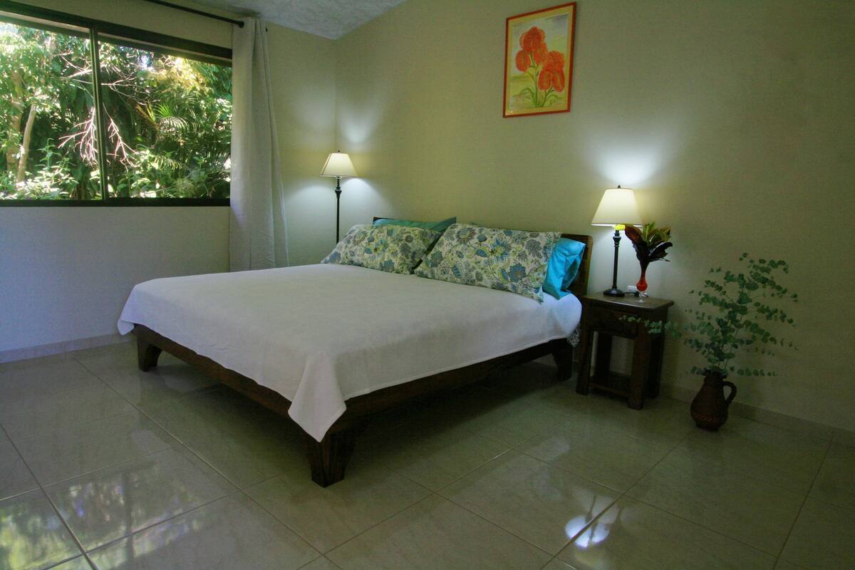 Un dormitorio con una cama, dos lámparas de noche, una planta en maceta y un ventanal con cortinas.