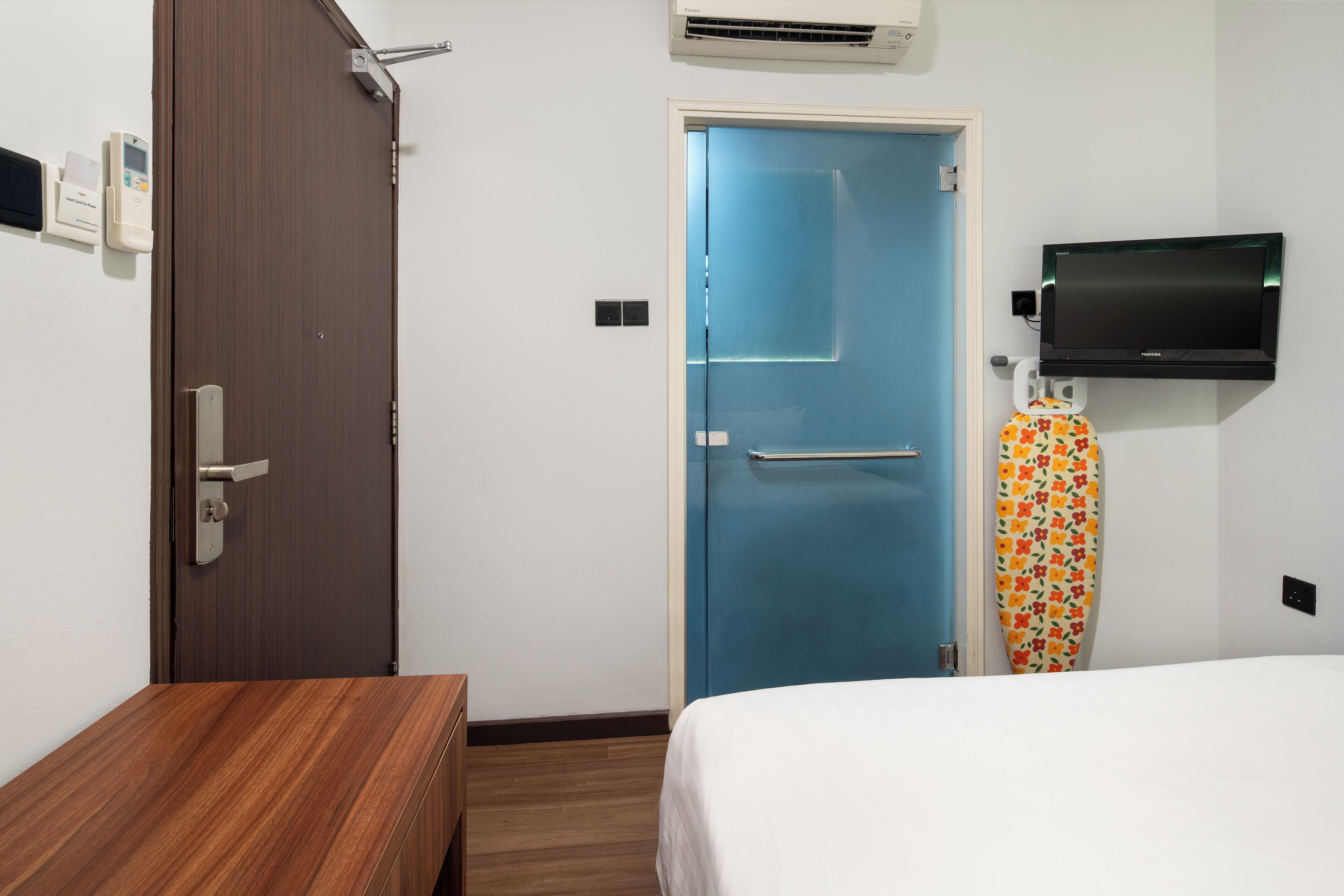 Une chambre d’hôtel avec une table de chevet en bois, un lit avec une literie blanche, une porte bleue, un téléviseur fixé au mur et un rideau de douche à motifs.