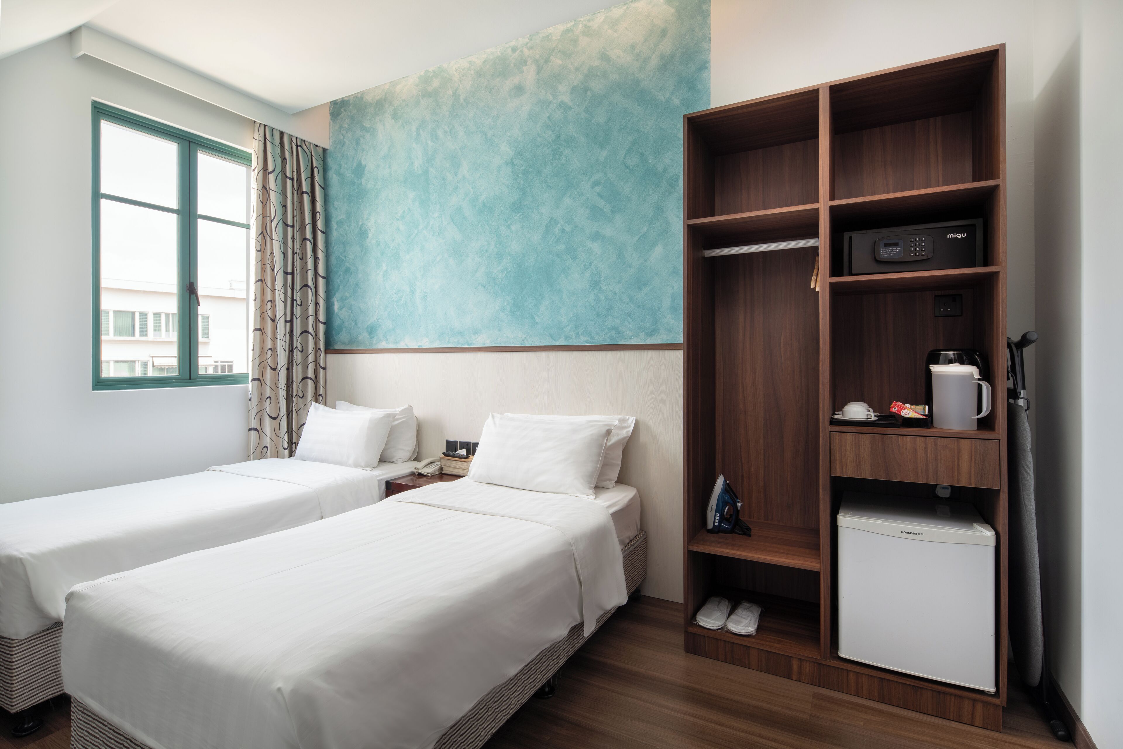 Una habitación de hotel con dos camas, un armario, un refrigerador pequeño y una ventana con cortinas.