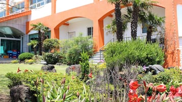 A building with orange and white exterior walls, a covered outdoor area with palm trees, and a garden with various plants and flowers.