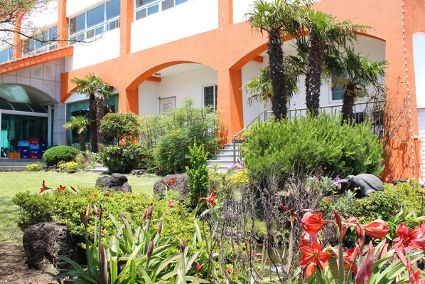 A building with orange and white exterior walls, a covered outdoor area with palm trees, and a garden with various plants and flowers. - Jeju Feel House (Jeju City)