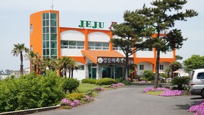 Un edificio con un llamativo letrero que dice "JEJU", rodeado de jardines cuidados y un camino de entrada despejado.