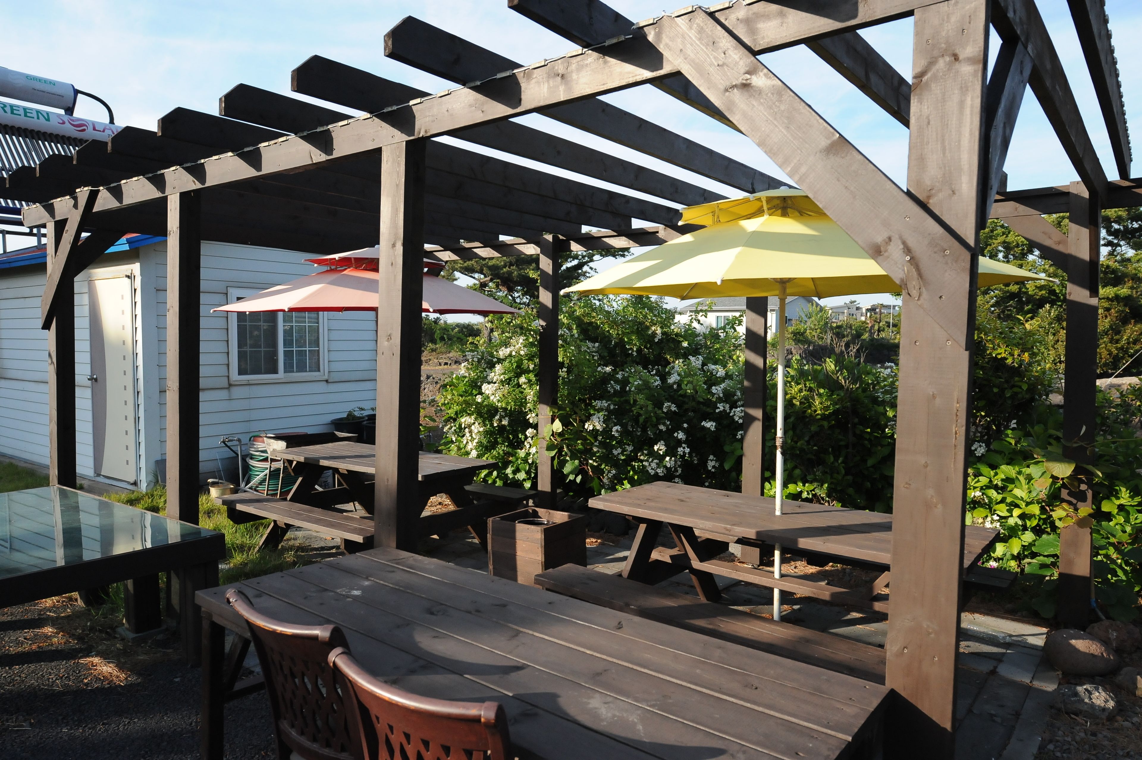 Eine Holz-Pergola mit Picknicktischen und Sonnenschirmen, umgeben von viel Grün und einem weißen Gebäude.