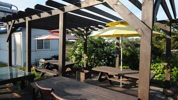 Eine Holz-Pergola mit Picknicktischen und Sonnenschirmen, umgeben von viel Grün und einem weißen Gebäude.