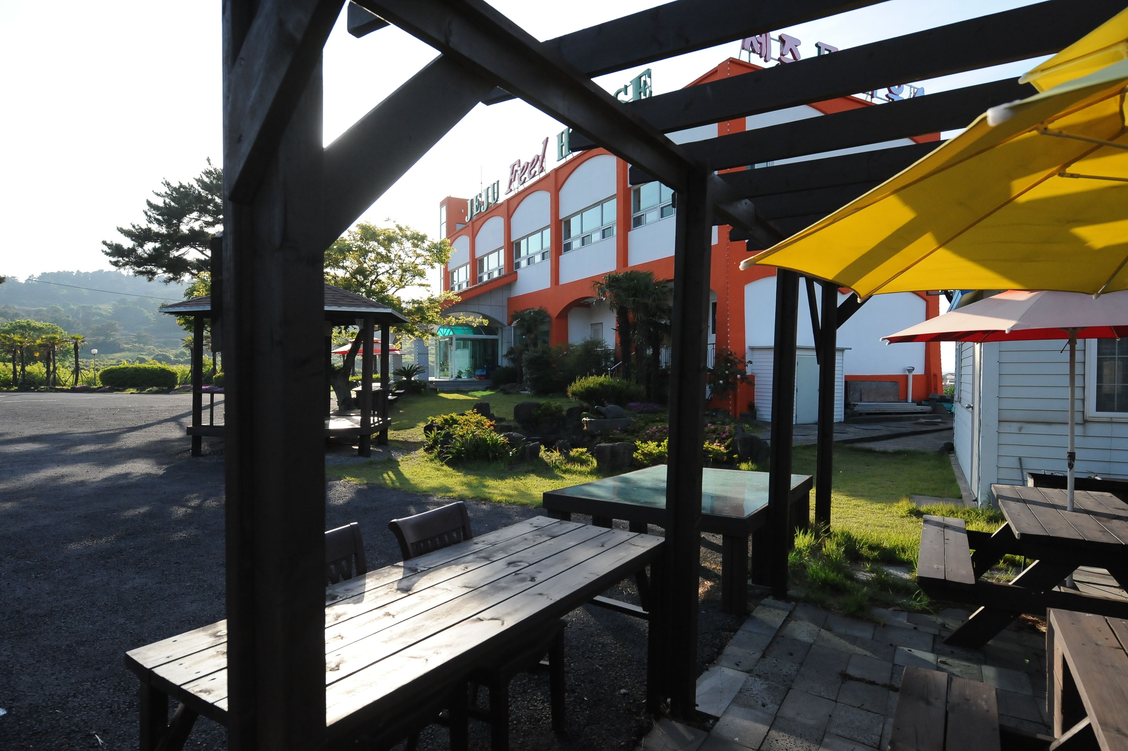 An outdoor patio area with wooden tables and benches, a yellow umbrella, and a building with a red and white facade in the background.