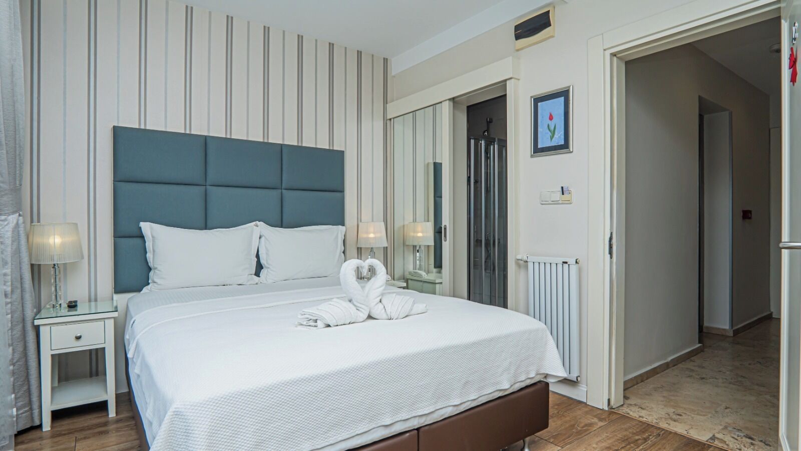 A hotel room with a large bed, a headboard, two bedside tables with lamps, a radiator, and a door leading to another room.
