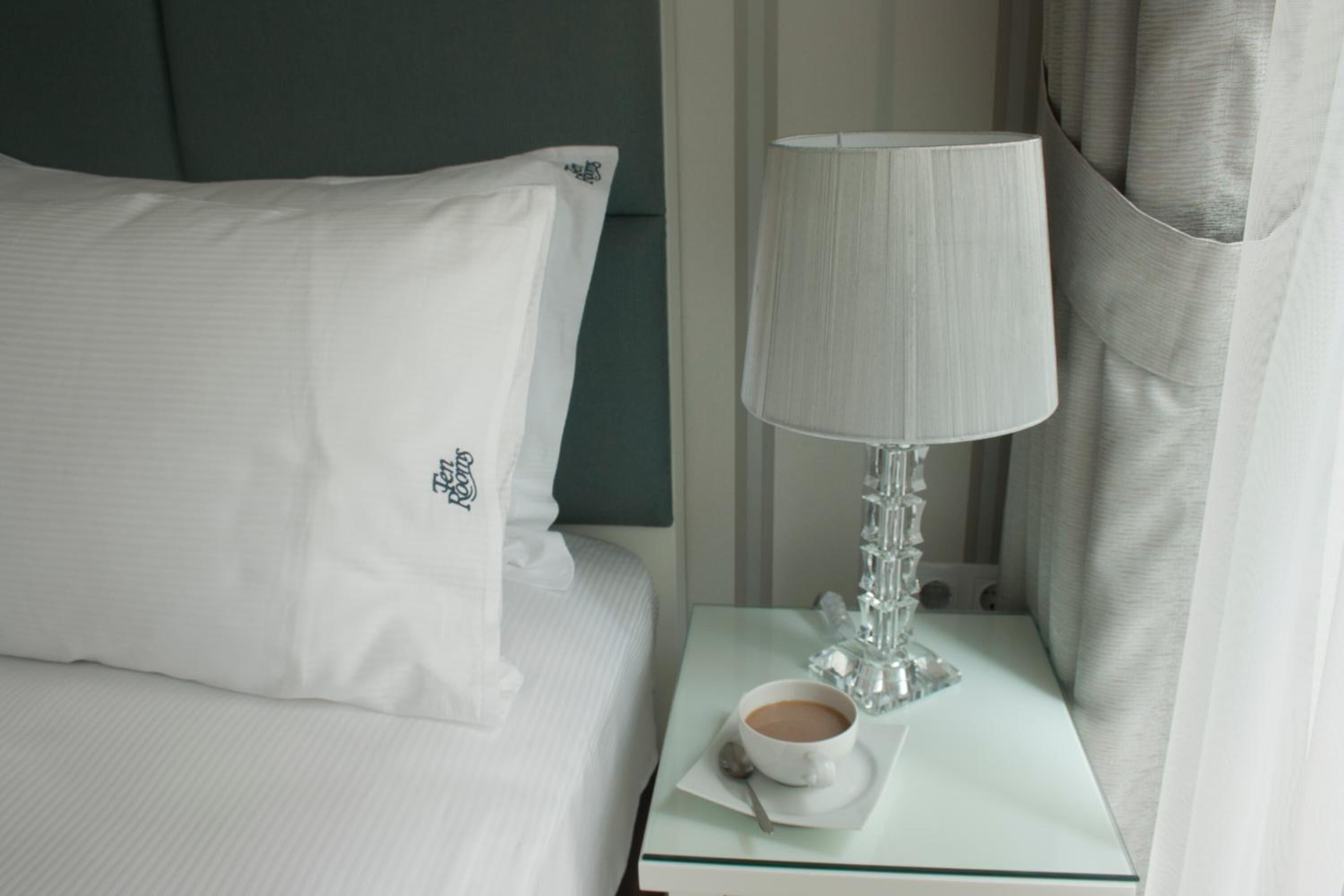 A neatly made bed with a white pillow and a glass table beside it, holding a cup of coffee and a glass.