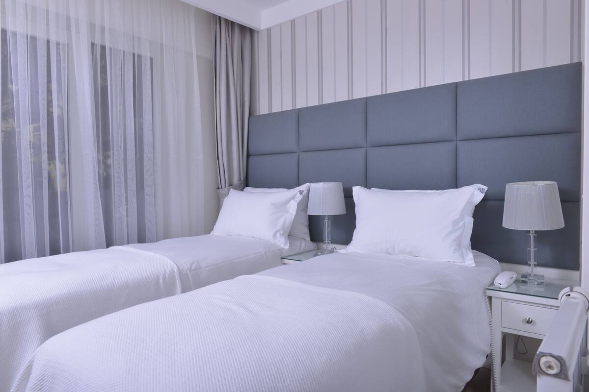 A hotel room with two beds, a grey headboard, white bedding, a bedside table with a lamp, and a phone.