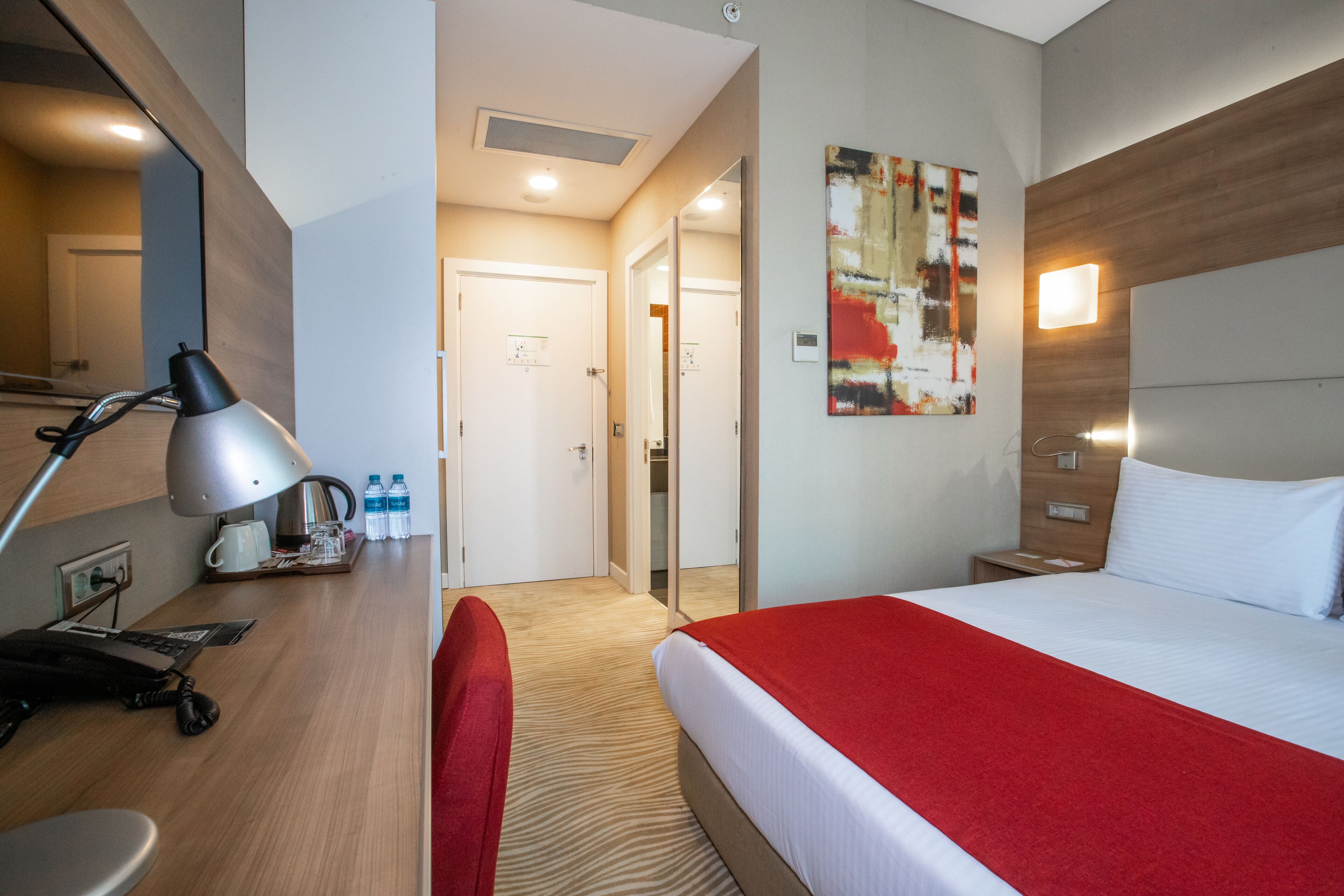 A hotel room with a bed, a desk with a lamp, a chair, a mirror, a red blanket, a wall art piece, and a door leading to a bathroom.