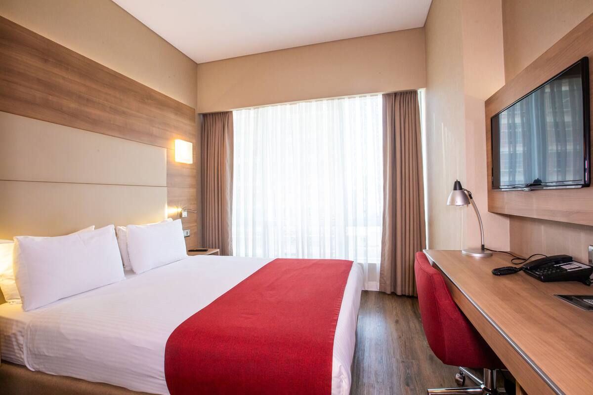 A hotel room with a large bed, a desk with a chair, a television, and a window with curtains.