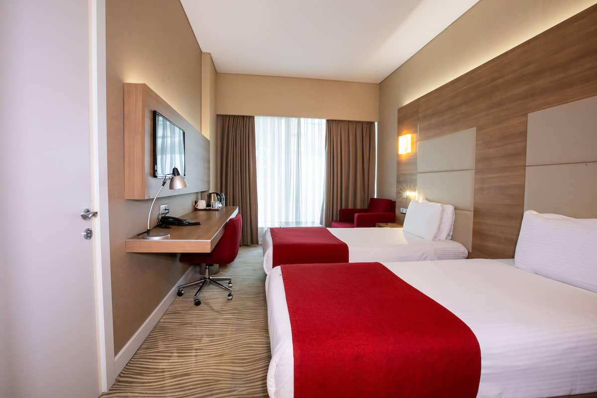 A hotel room with two beds, a desk, a chair, and a large window with curtains.
