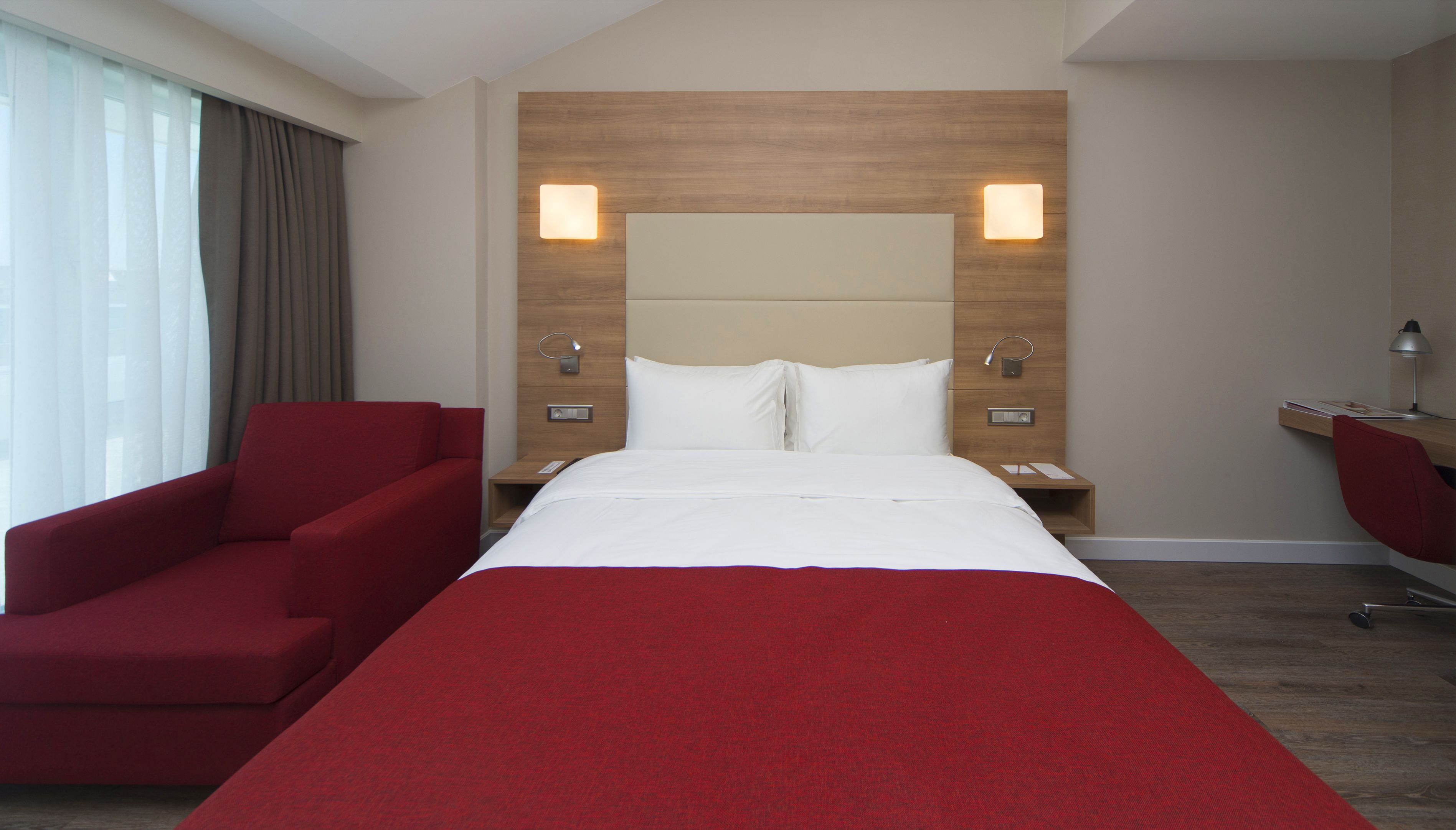 Ein Hotelzimmer mit einem großen Bett, einem roten Sessel, einem Schreibtisch mit Lampe und einem Nachttisch ebenfalls mit Lampe.