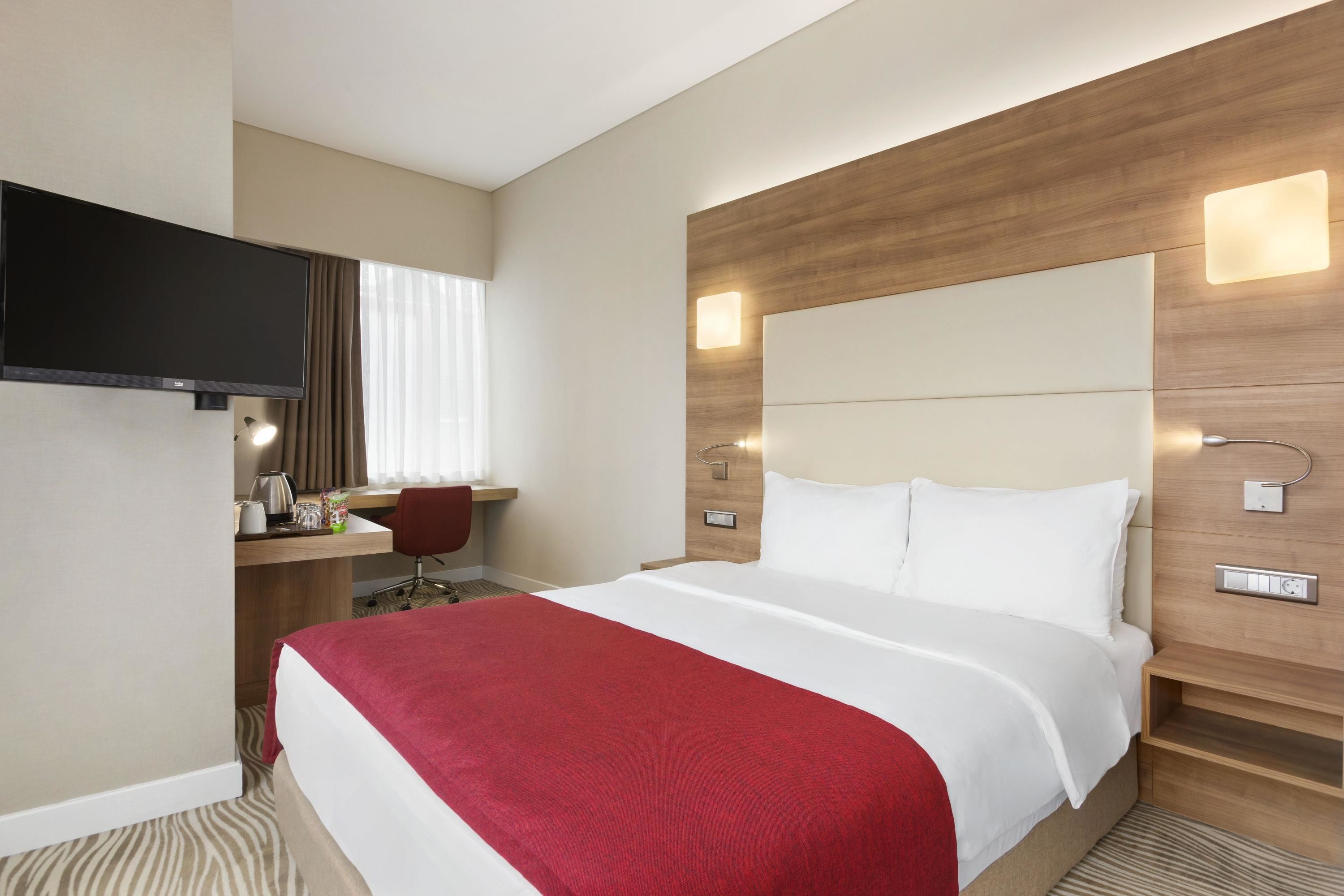 A hotel room with a large bed, a desk with a chair, a flat-screen TV, and a wooden headboard with built-in lighting.