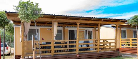 A mobile home with a wooden deck, a covered porch, and a small tree.