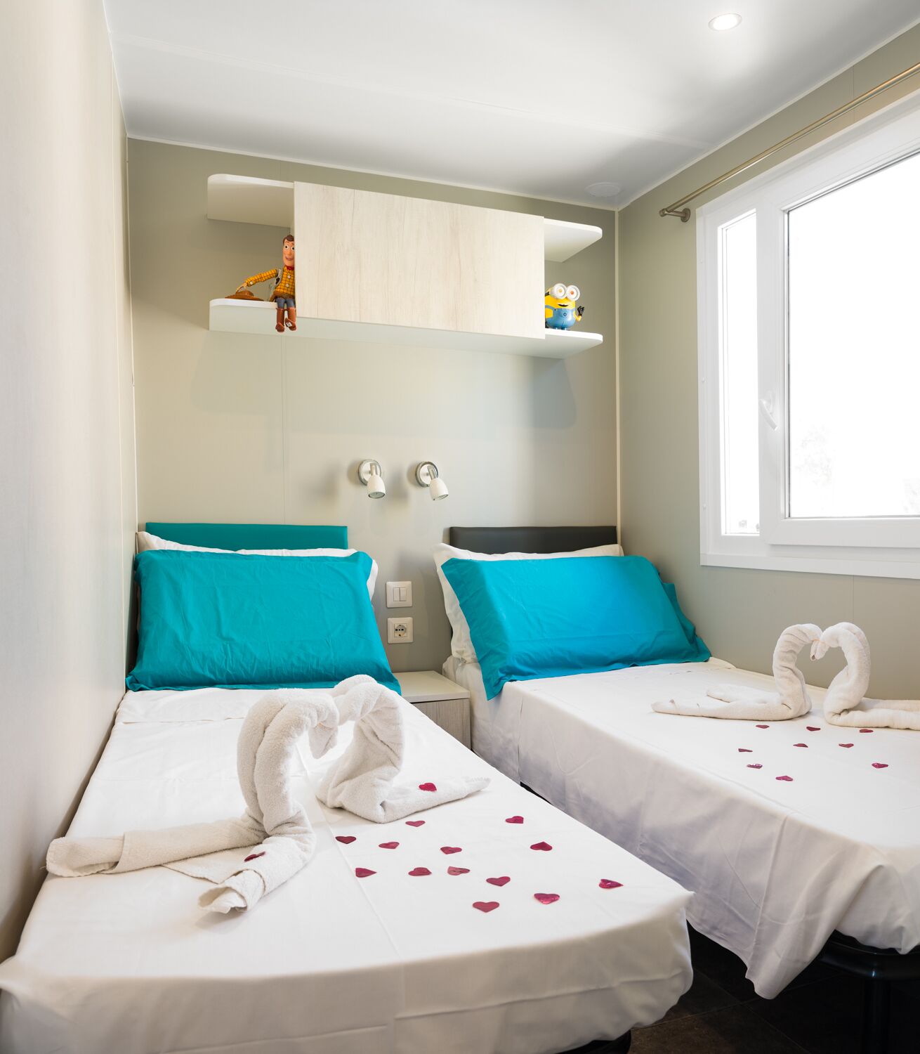 Una habitación de hotel con dos camas individuales, cada una con una almohada color turquesa y una manta blanca. Hay dos toallas blancas sobre las camas, y un par de toallas con forma de cisne en el alféizar.